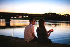 Boy and a girl holding a sprinkler sitting together at the riverbank with a bridge in the background at sunset.
