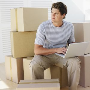 A man sitting on a pile of boxes with his laptop, looking away from it, trying to figure out when's the cheapest time to relocate