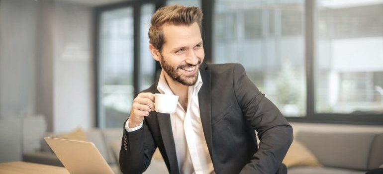 A man drinking a cup of coffee at his desk and smiling.