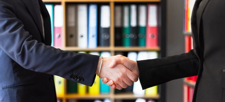 Two people in suits shaking hands with a bookshelf in the background.