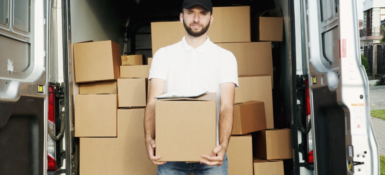 A man in a white shirt carrying a cardboard box in front of other boxes.