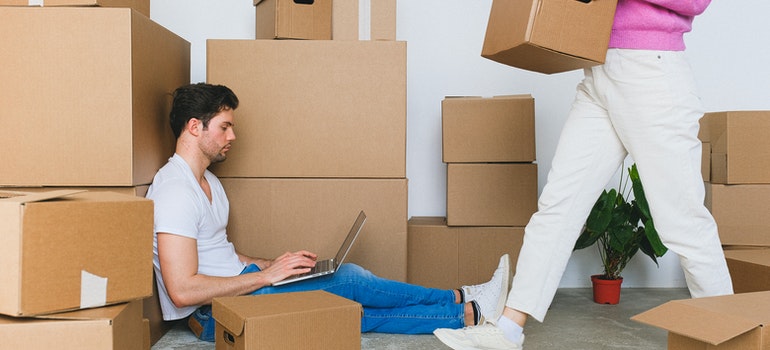 A man sitting among moving boxes and a woman walking by carrying a box