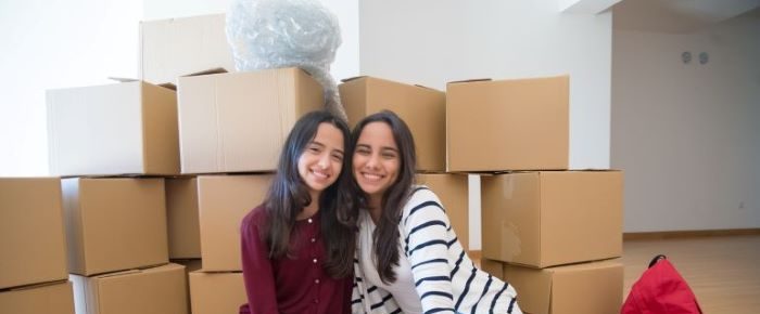 Two girls sitting and smiling in front of a pile of moving boxes.